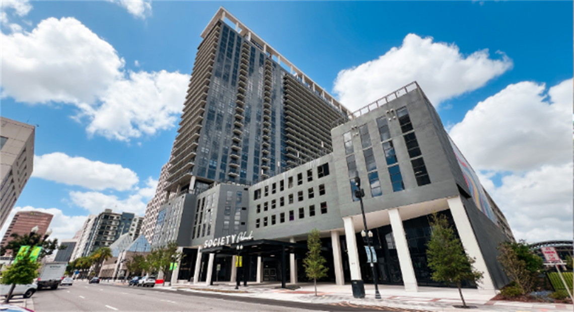 Tall mixed‑use building with large windows and commercial space at the base under a blue sky.