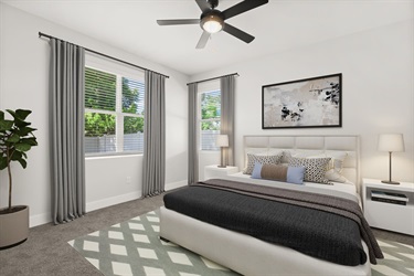 Bright bedroom with a large bed, white upholstered headboard, and layered bedding in neutral tones. Two tall windows with gray curtains flank the bed, and a ceiling fan hangs above. A patterned rug and a potted plant complete the modern, clean design.