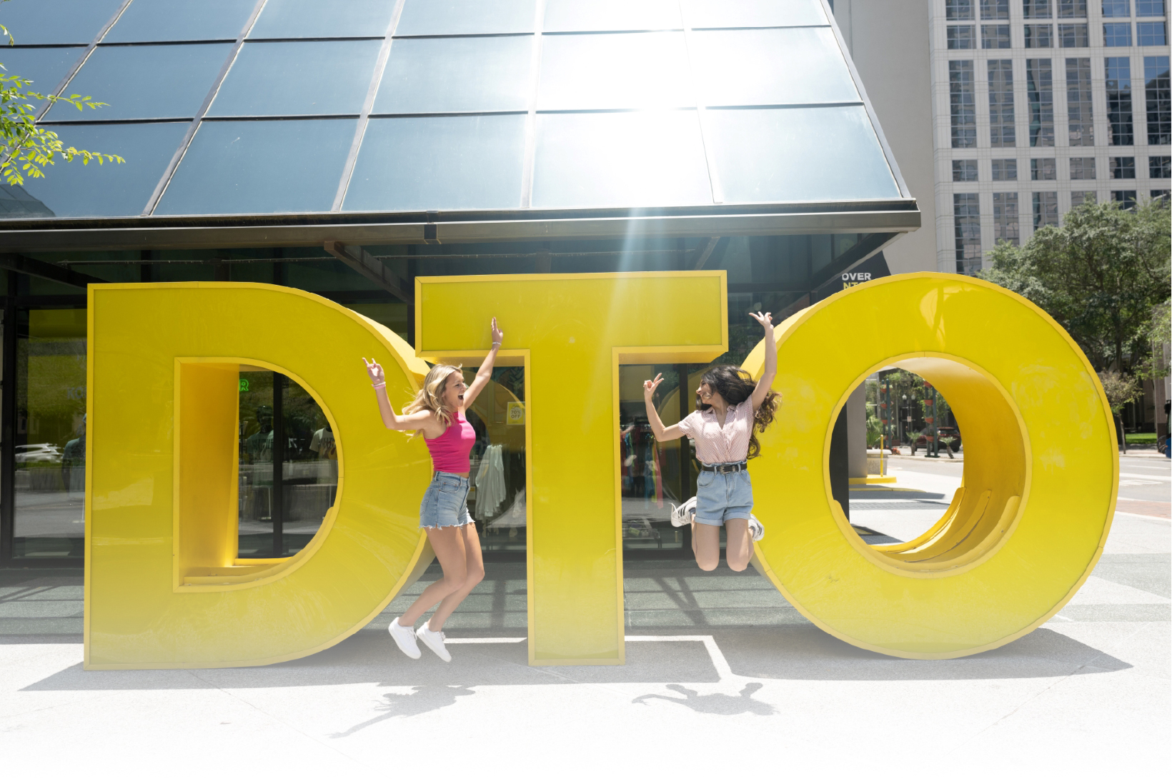 Two girls smiling and jumping in front of a large yellow DTO sculpture