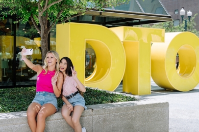 Two women posing for a selfie in front of a large yellow DTO sculpture