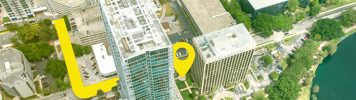 An aerial view of a city block with tall office buildings, roads, trees, and a nearby body of water. A large yellow graphic path curves through the scene, ending in a yellow location pin symbol placed near the center of the buildings.